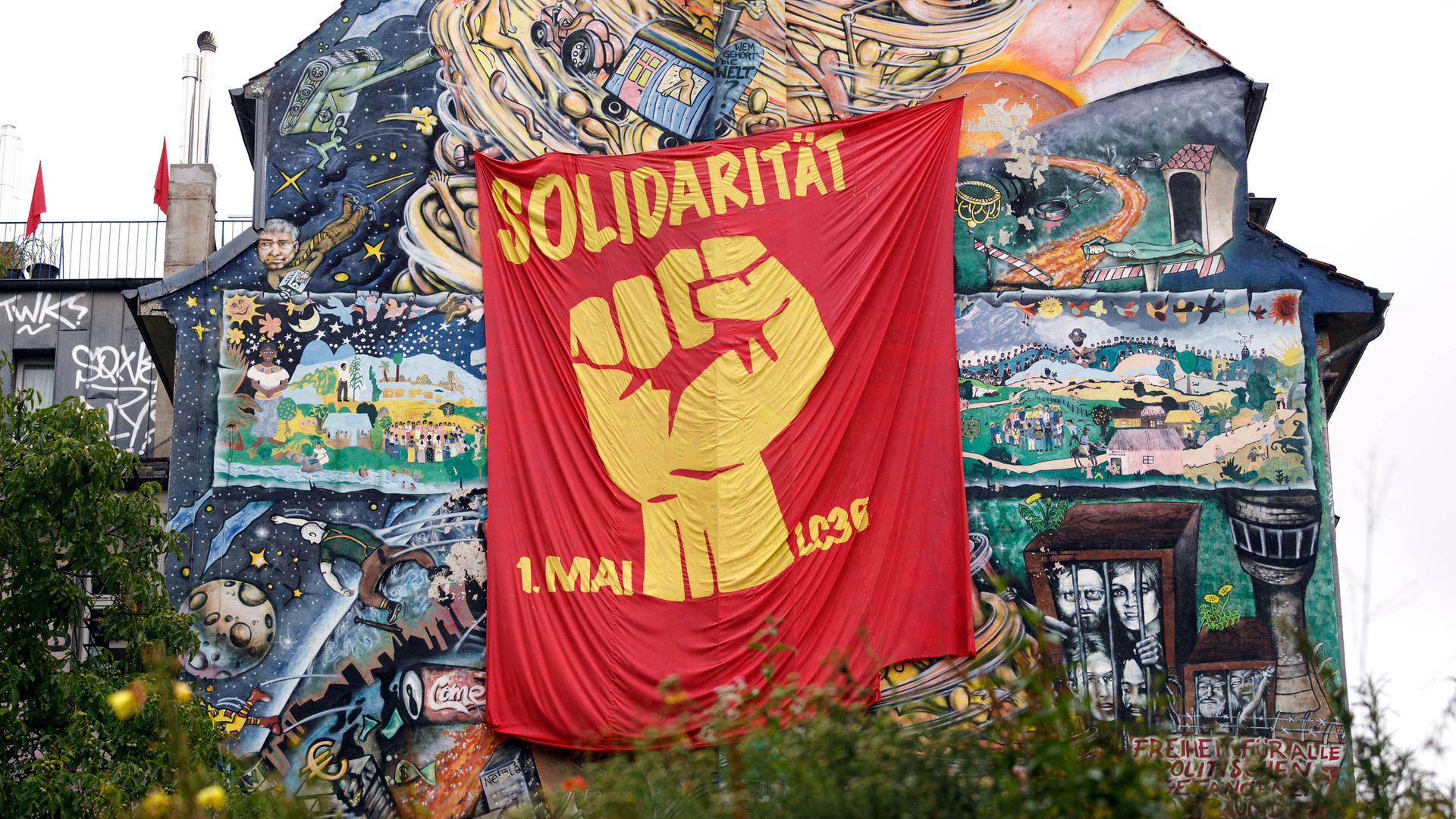 An einer hohen Hauswand, die mit zahlreichen Szenen bemalt wurde, hängt ein rotes Tuch. Darauf sieht man eine geballte Faust und die Worte "Solidarität 1. Mai". An einer hohen Hauswand, die mit zahlreichen Szenen bemalt wurde, hängt ein rotes Tuch. Darauf sieht man eine geballte Faust und die Worte "Solidarität 1. Mai".