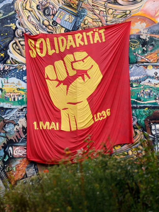 An einer hohen Hauswand, die mit zahlreichen Szenen bemalt wurde, hängt ein rotes Tuch. Darauf sieht man eine geballte Faust und die Worte "Solidarität 1. Mai".