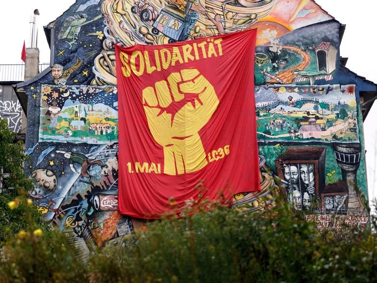 An einer hohen Hauswand, die mit zahlreichen Szenen bemalt wurde, hängt ein rotes Tuch. Darauf sieht man eine geballte Faust und die Worte "Solidarität 1. Mai".