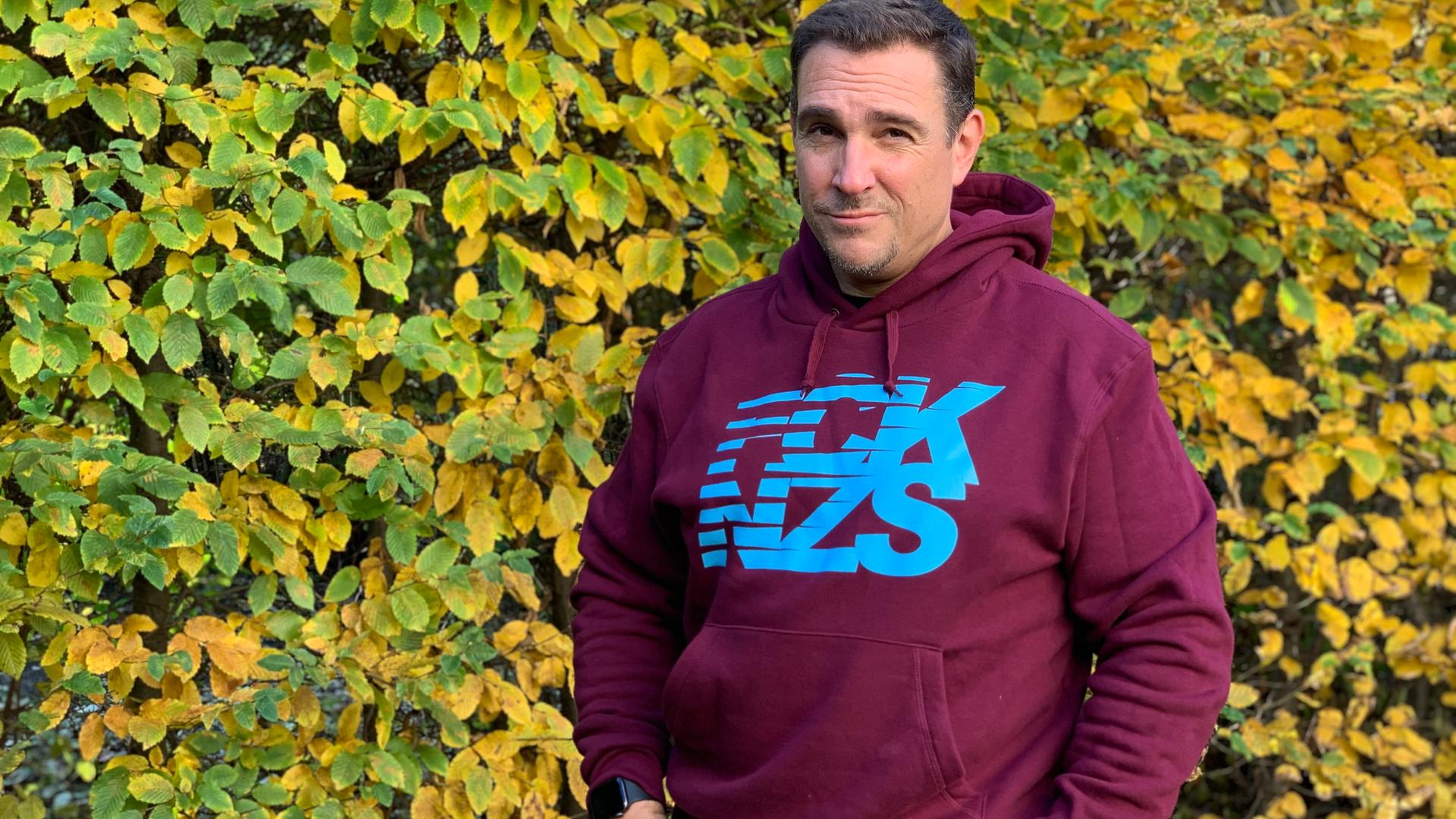 Oliver von Dobrowolski steht mit einem FCK NZS Pullover vor einer Hecke und schaut in die Kamera. Oliver von Dobrowolski steht mit einem FCK NZS Pullover vor einer Hecke und schaut in die Kamera.