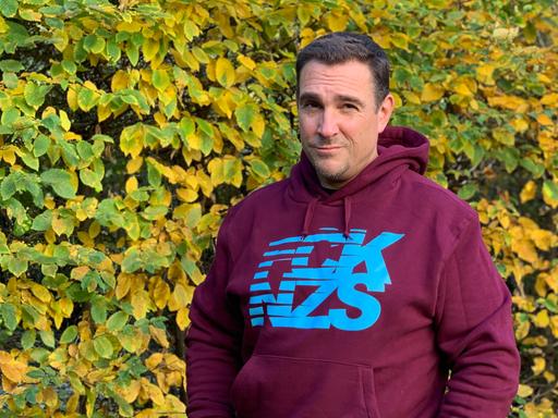 Oliver von Dobrowolski steht mit einem FCK NZS Pullover vor einer Hecke und schaut in die Kamera.