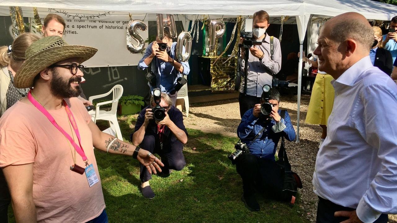 links steht Anas Aboura mit einem Strohhut und reicht dem auf der rechten Bildseite stehendem Olaf Scholz die Hand zum Gruß hin. Im Hintergrund sind einige Fotografen zu sehen. links steht Anas Aboura mit einem Strohhut und reicht dem auf der rechten Bildseite stehendem Olaf Scholz die Hand zum Gruß hin. Im Hintergrund sind einige Fotografen zu sehen.