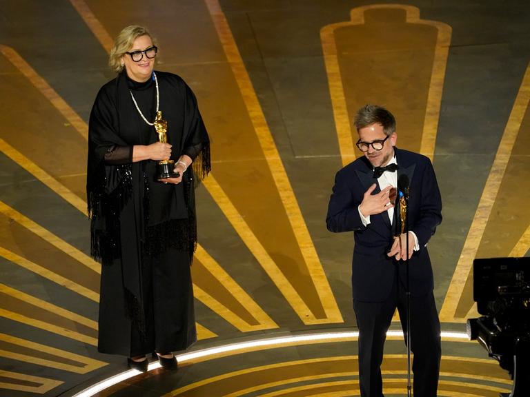 Ernestine Hipper im schwarzen Kleid und Christian M. Goldbeck im Smoking halten jeweils eine goldene Oscar-Statue in Händen.