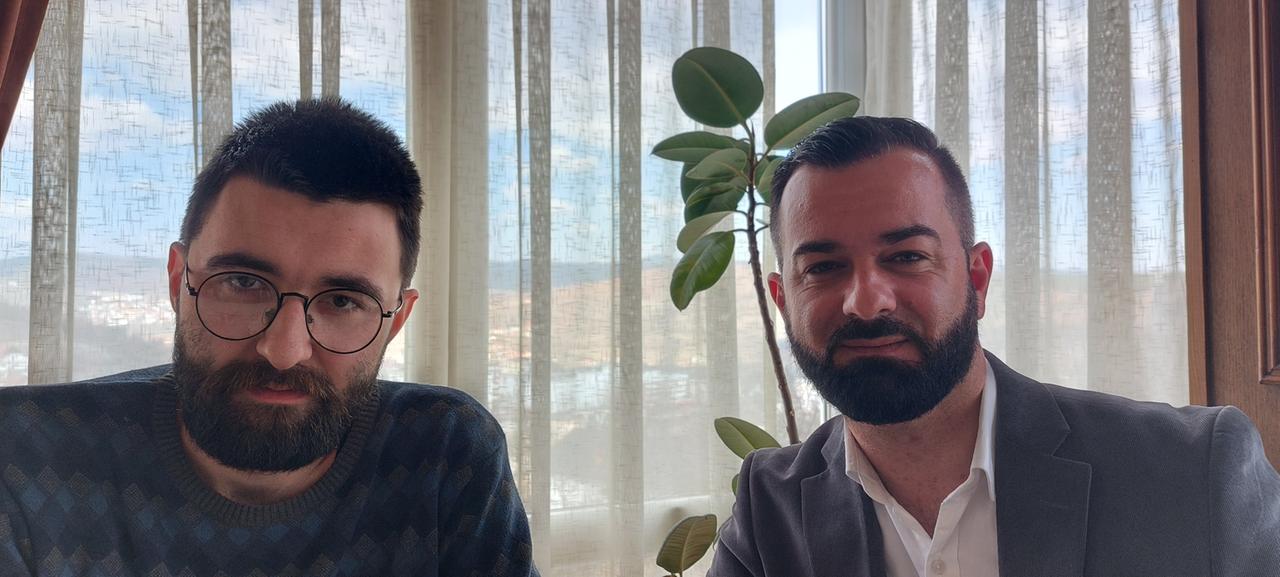 Zwei mittelalte Männer mit kurzen dunklen Haaren und Bart sitzen vor einem Vorhang und gucken in die Kamera Zwei mittelalte Männer mit kurzen dunklen Haaren und Bart sitzen vor einem Vorhang und gucken in die Kamera