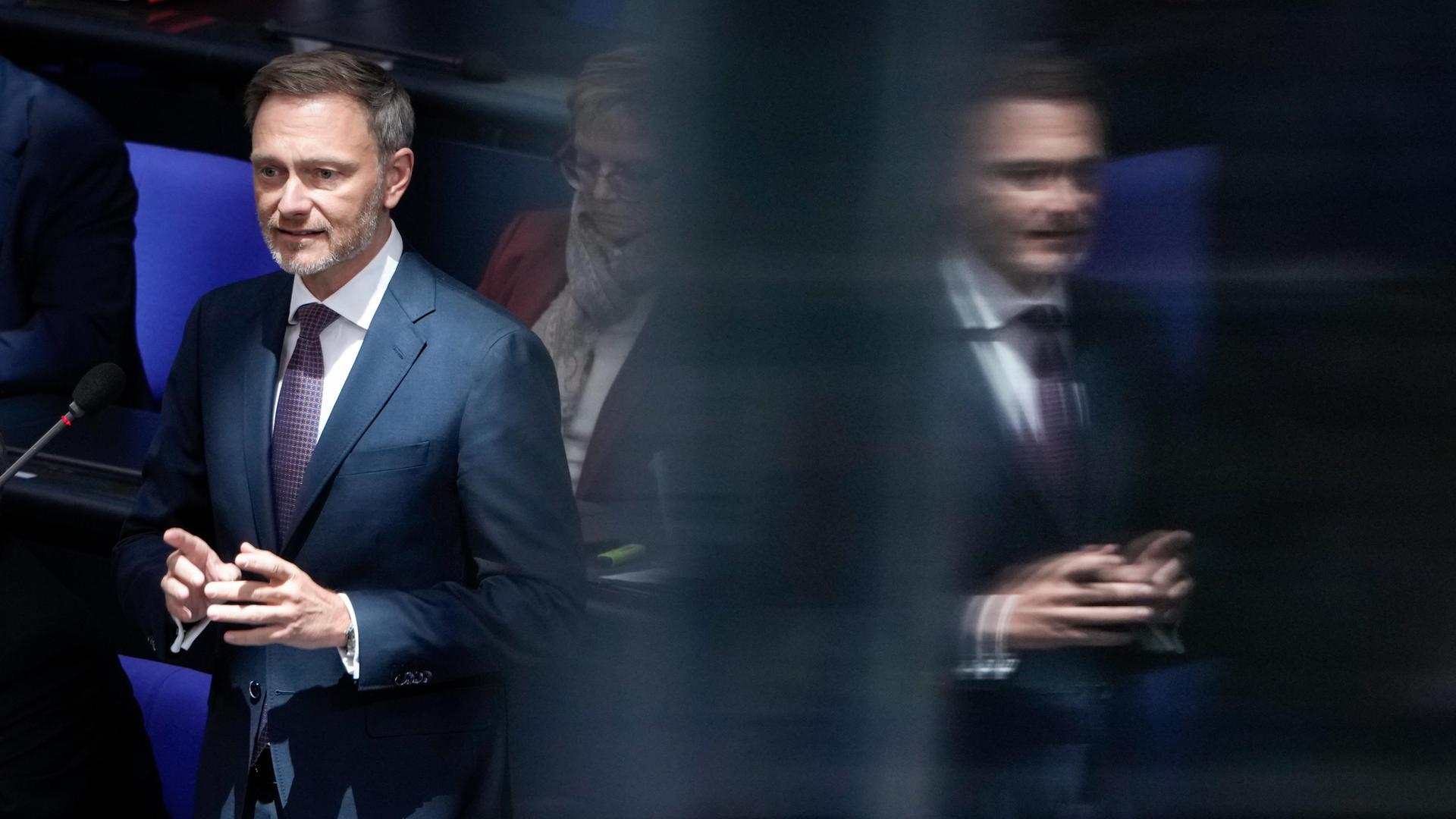 Porträt des Bundesfinanzministers Christian Lindner (FDP): Er steht an seinem Platz auf der Regierungsbank vor einem Mikrofon und spiegelt sich rechts daneben in einer Scheibe. Porträt des Bundesfinanzministers Christian Lindner (FDP): Er steht an seinem Platz auf der Regierungsbank vor einem Mikrofon und spiegelt sich rechts daneben in einer Scheibe.