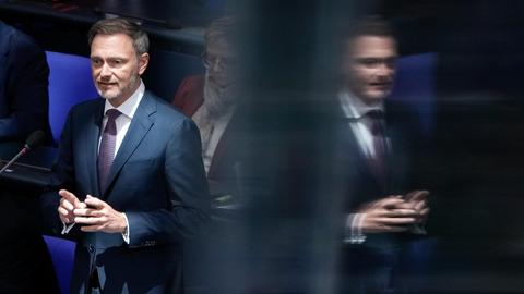 Porträt des Bundesfinanzministers Christian Lindner (FDP): Er steht an seinem Platz auf der Regierungsbank vor einem Mikrofon und spiegelt sich rechts daneben in einer Scheibe.