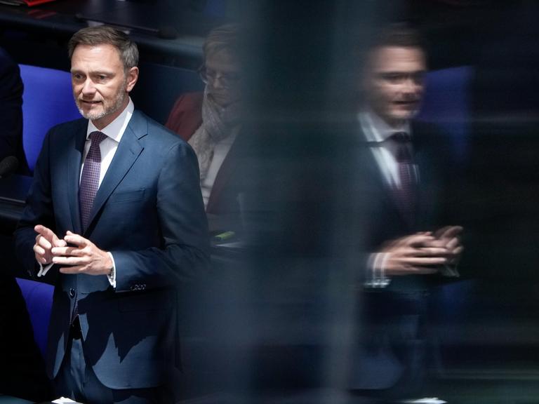 Porträt des Bundesfinanzministers Christian Lindner (FDP): Er steht an seinem Platz auf der Regierungsbank vor einem Mikrofon und spiegelt sich rechts daneben in einer Scheibe.