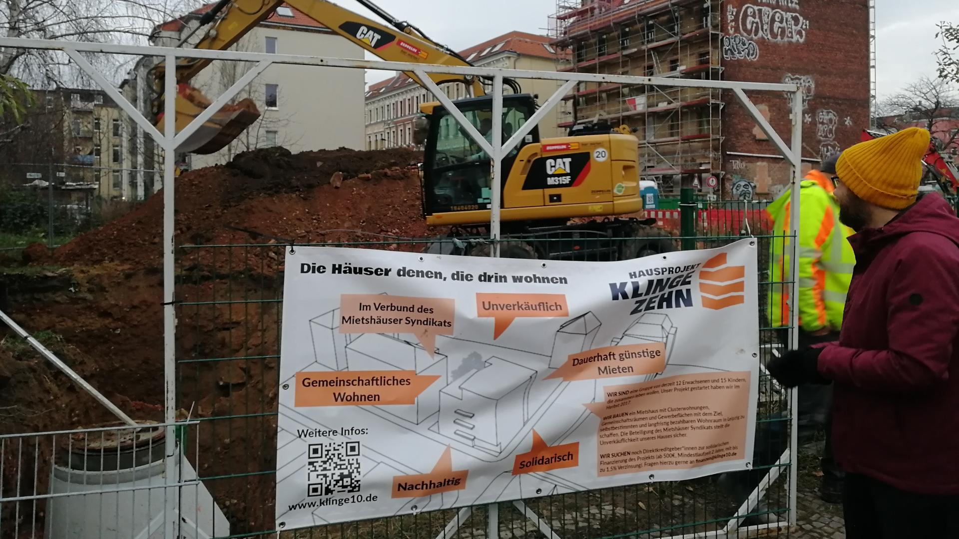 Ein Bagger auf einer Baustelle. Auf dem Bauzaun steht "die Häuser denen, die darin Wohnen Ein Bagger auf einer Baustelle. Auf dem Bauzaun steht "die Häuser denen, die darin Wohnen