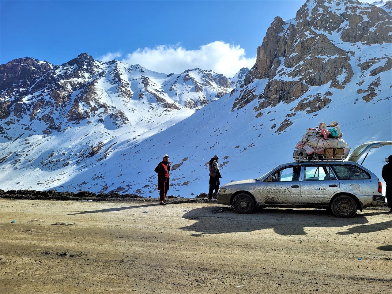 Auf einer Gebirgsstraße im schneebedeckten Hindukusch steht ein altes Auto mit viel Gepäck auf dem Dach, das von Taliban kontrolliert wird.
