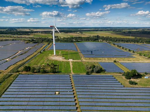 Eine Photovoltaik-Freiflächenanlage (Solarpark), betrieben von der Firma Enerparc. Zahlreiche Solarzellen in dem Solarkraftwerk wandeln Sonnenenergie in grünen Strom um. In dem Solarpark steht ein Windrad. (Luftaufnahme mit Drohne)