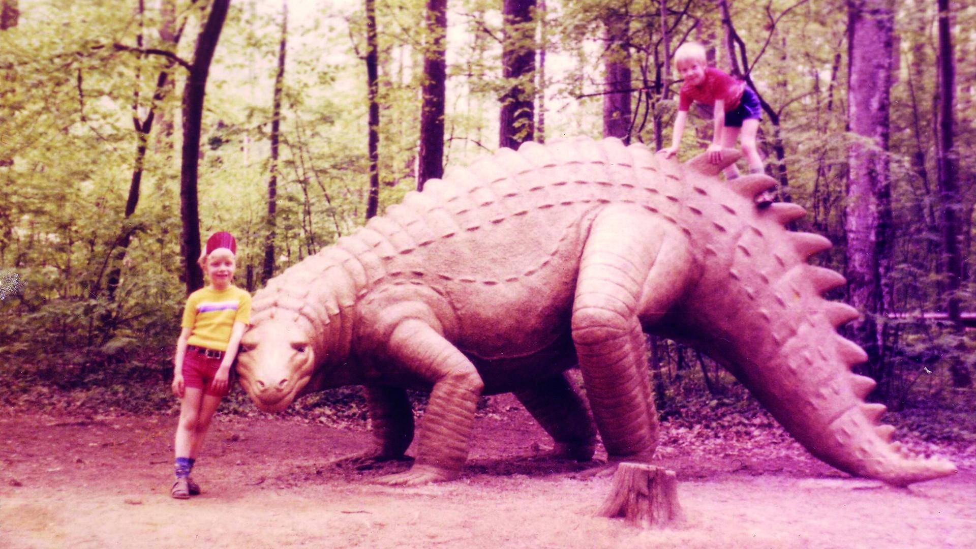Zwei Kinder posieren neben dem lebensgroßen Modell eines Dinosauriers in einem Freizeitpark. Zwei Kinder posieren neben dem lebensgroßen Modell eines Dinosauriers in einem Freizeitpark.