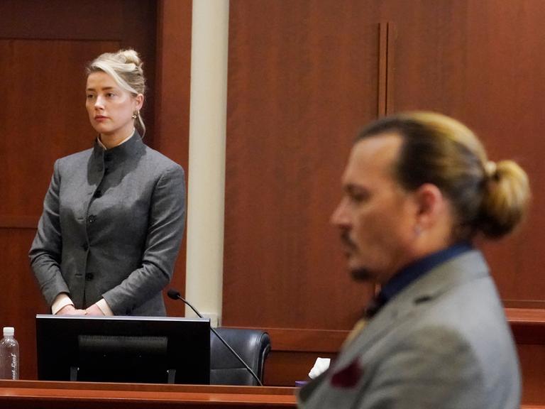 Amber Heard stehend bei einer Aussage im Gerichtssaal, im Vordergrund hört ihr Ex-Ehemann Johnny Depp zu.