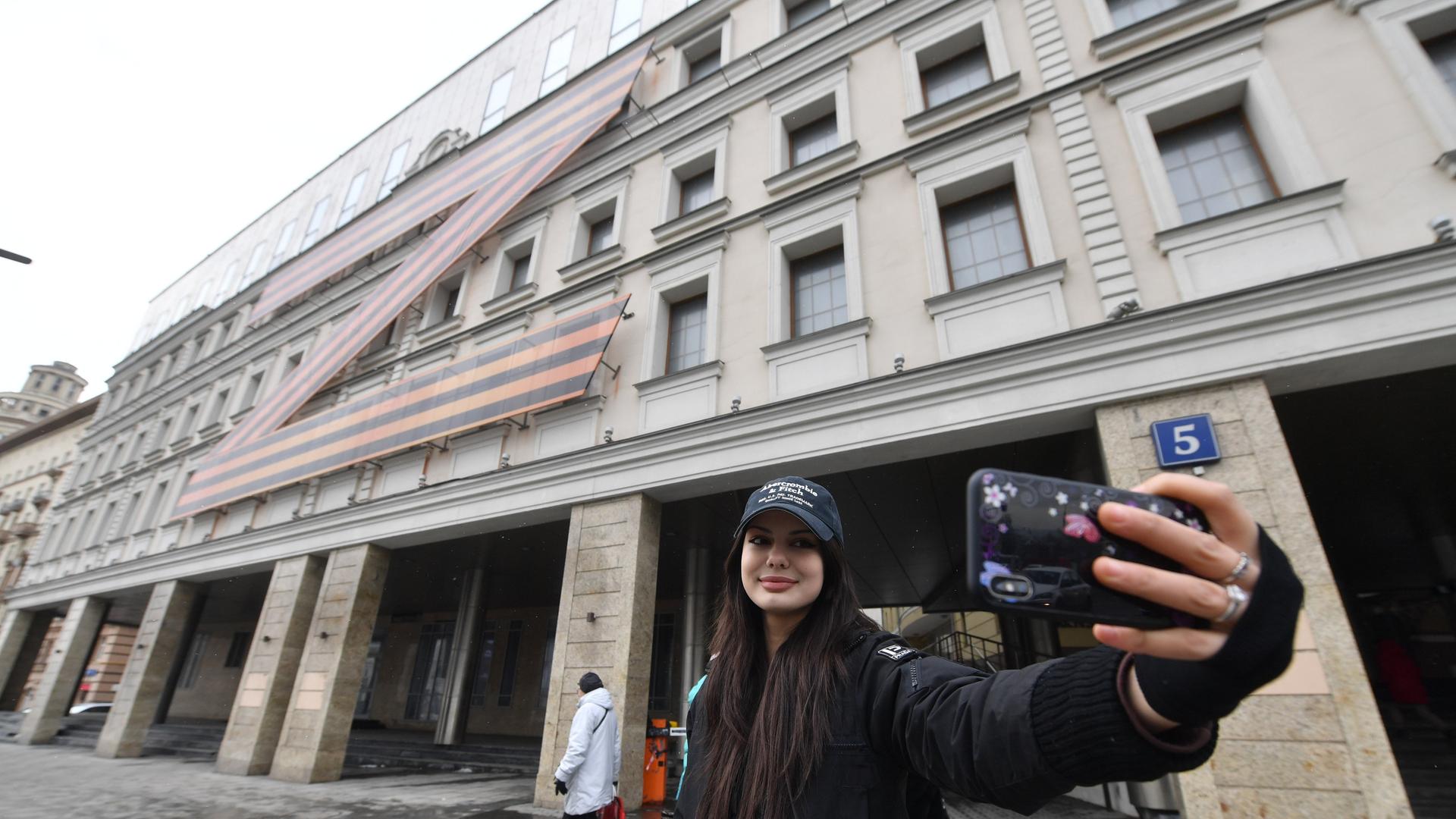 Eine junge Frau fotografiert sich vor einem Gebäude in Moskau, auf dem das Z-Symbol zu sehen ist. Eine junge Frau fotografiert sich vor einem Gebäude in Moskau, auf dem das Z-Symbol zu sehen ist.
