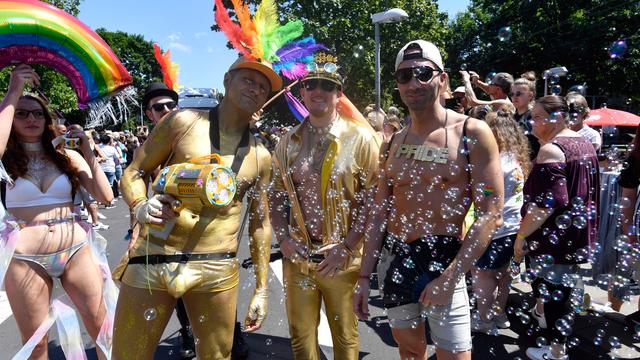 Drei Männer mit nacktem Oberkörper stehen auf einer Straße, zwei von ihen sind bronzefarben angemalt. Einer trägt einen Hut mit Federn in den Regenbogenfarben. Drumnherum weitere Teilnehmer.