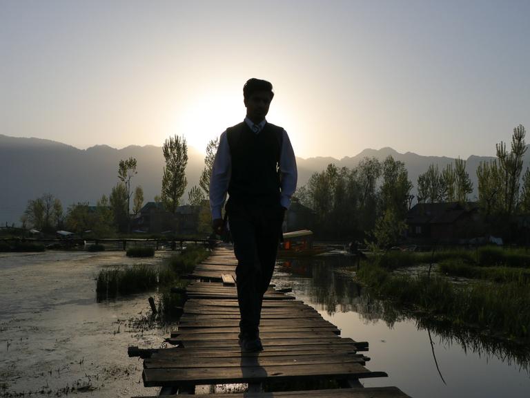 Silhouette eines gut gekleideten Mannes, der geradeaus auf einem Steg entlang läuft. Rechts und links davon ist Wasser. Im Hintergrund sind Berge zu erkennen.