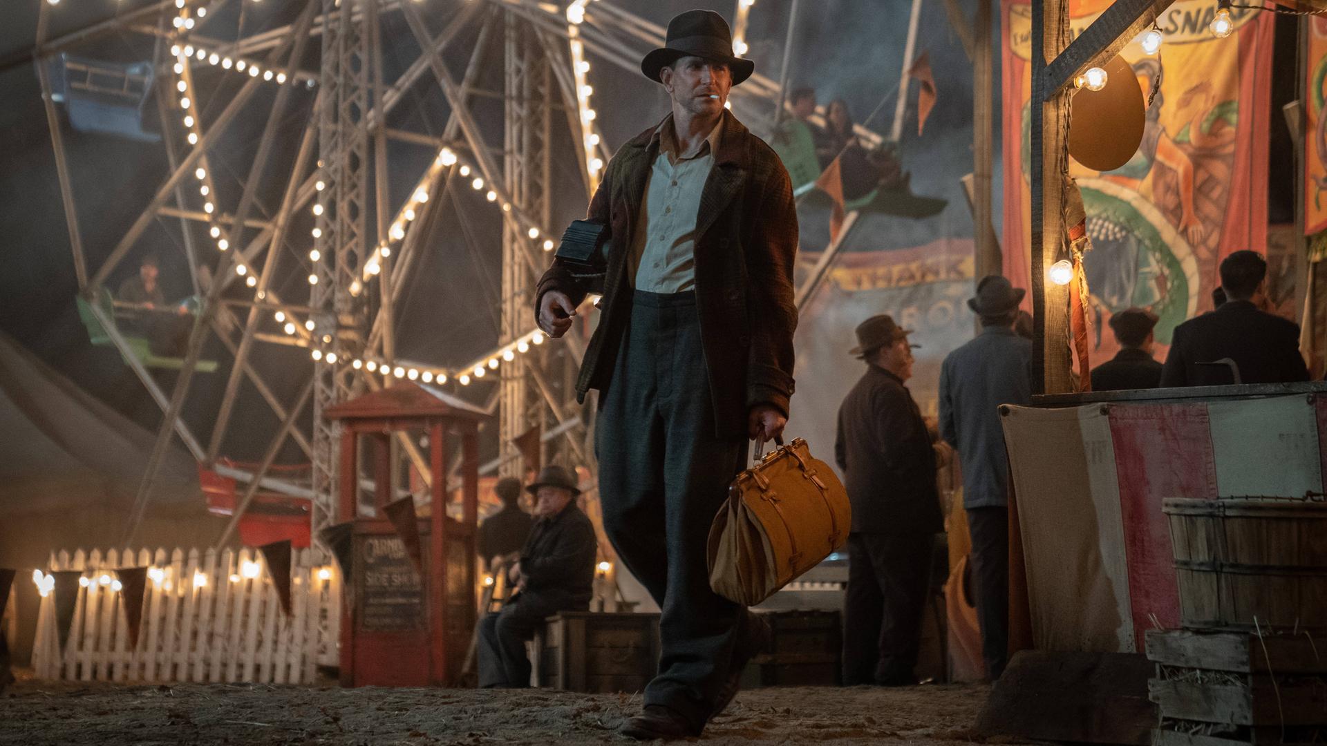 In dem Film Nightmare Alley von Regisseur Guillermo del Toro geht der von Bradley Cooper gespielte Betrüger Stan Carlisle über einen von einem Riesenrad erleuchteten Jahrmarkt. In dem Film Nightmare Alley von Regisseur Guillermo del Toro geht der von Bradley Cooper gespielte Betrüger Stan Carlisle über einen von einem Riesenrad erleuchteten Jahrmarkt.