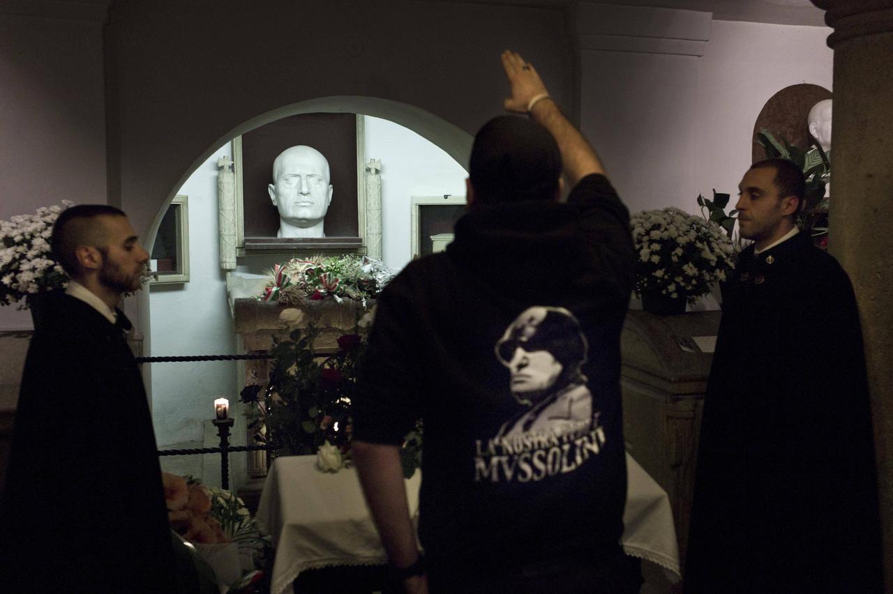 Ein Mann mit schwarzem Sweater, auf dessen Rückseite Mussolini abgebildet ist, streckt den gestreckten rechten Arm einer Mussolini-Büste an einer Gedenkstätte mit Blumen und Kerzen entgegen. Ein Mann mit schwarzem Sweater, auf dessen Rückseite Mussolini abgebildet ist, streckt den gestreckten rechten Arm einer Mussolini-Büste an einer Gedenkstätte mit Blumen und Kerzen entgegen.