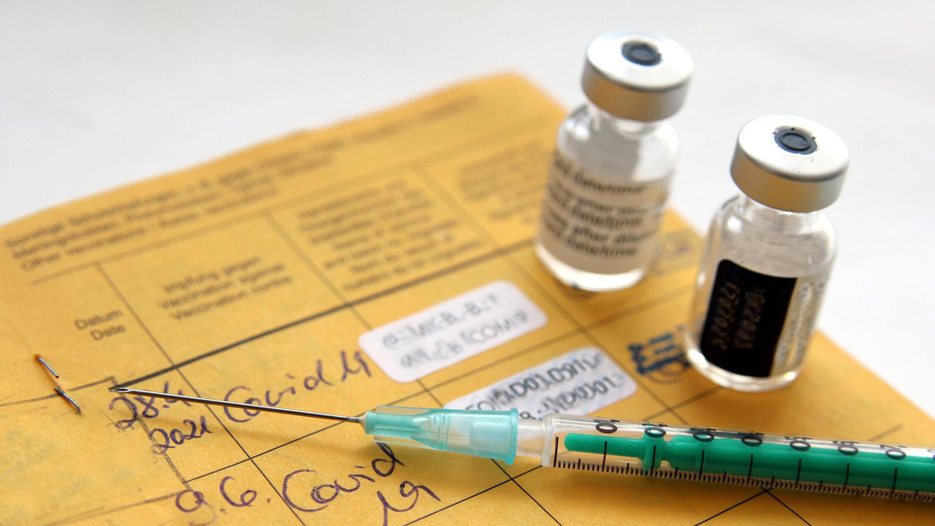 Impfdosen und eine Spritze liegen auf einem gelben Impfausweis.