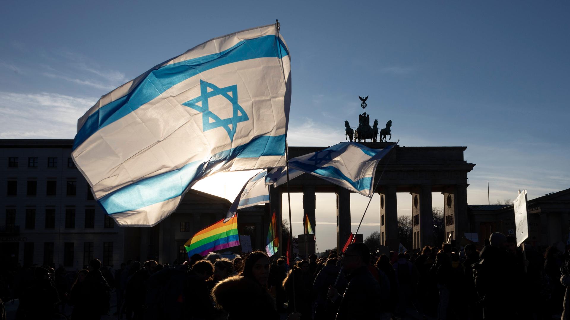 Eine israelische Fahne und eine Regenbogenfahne werden bei einer Demonstration geschwenkt. Im Hintergrund erkennt man das Brandenburger Tor in Berlin. Eine israelische Fahne und eine Regenbogenfahne werden bei einer Demonstration geschwenkt. Im Hintergrund erkennt man das Brandenburger Tor in Berlin.