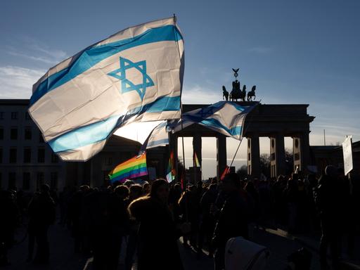 Eine israelische Fahne und eine Regenbogenfahne werden bei einer Demonstration geschwenkt. Im Hintergrund erkennt man das Brandenburger Tor in Berlin.