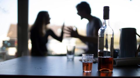 Ein Mann und eine Frau diskutieren heftig miteinander, im Vordergrund steht eine Flasche mit Alkohol auf dem Tisch
