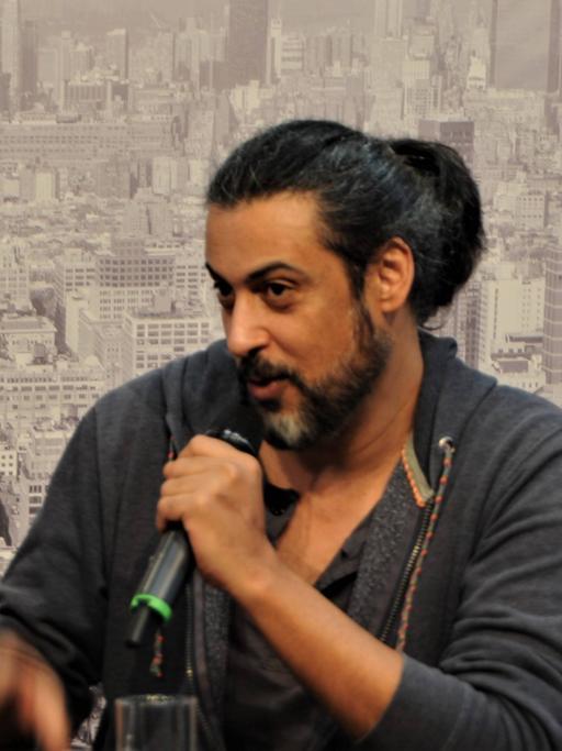 Abbas Khider hat dunkle Haare, die er zu einem Dutt zusammengebunden hat und trägt Bart. Im Hintergrund ist das Foto einer Stadtlandschaft zu sehen mit vielen Hochhäusern 