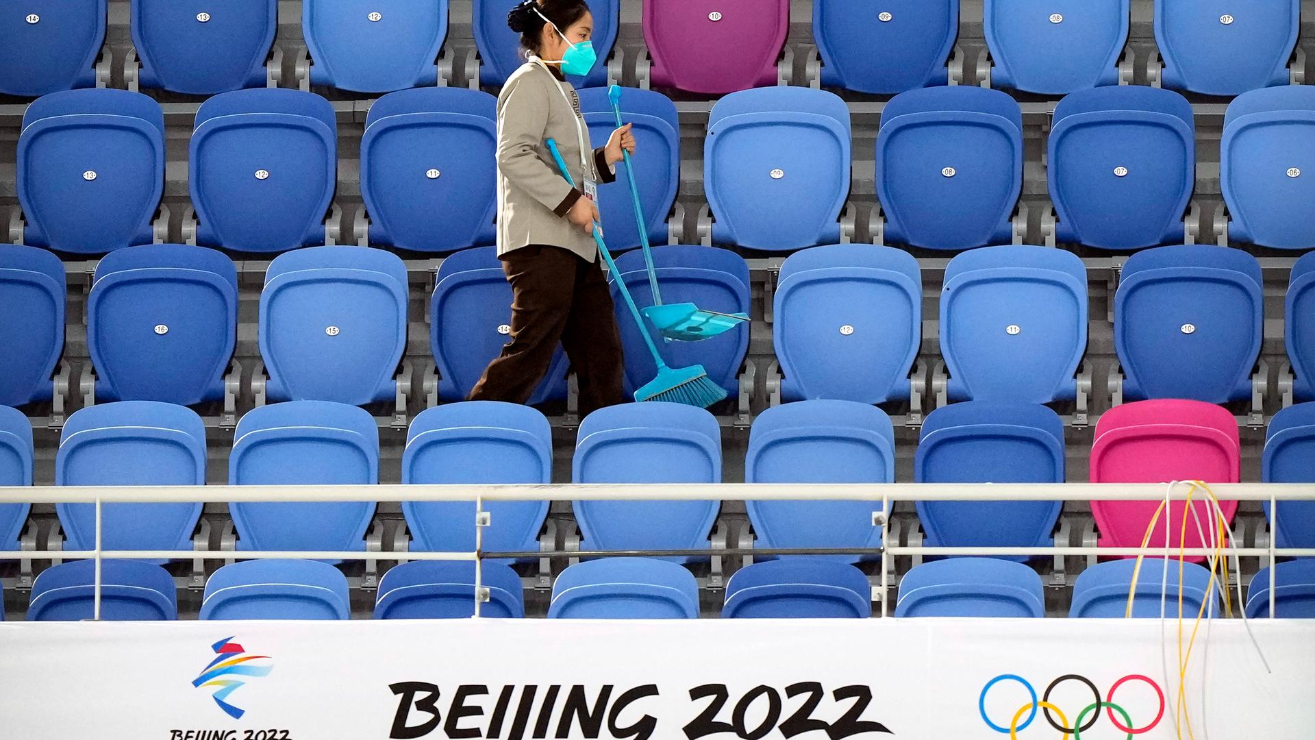Eine Frau mit Maske reinigt leere Ränge im Eisschnelllaufstadion von Peking. Vor ihr ist ein Logo der Olympischen Winterspiele 2022 zu sehen. 