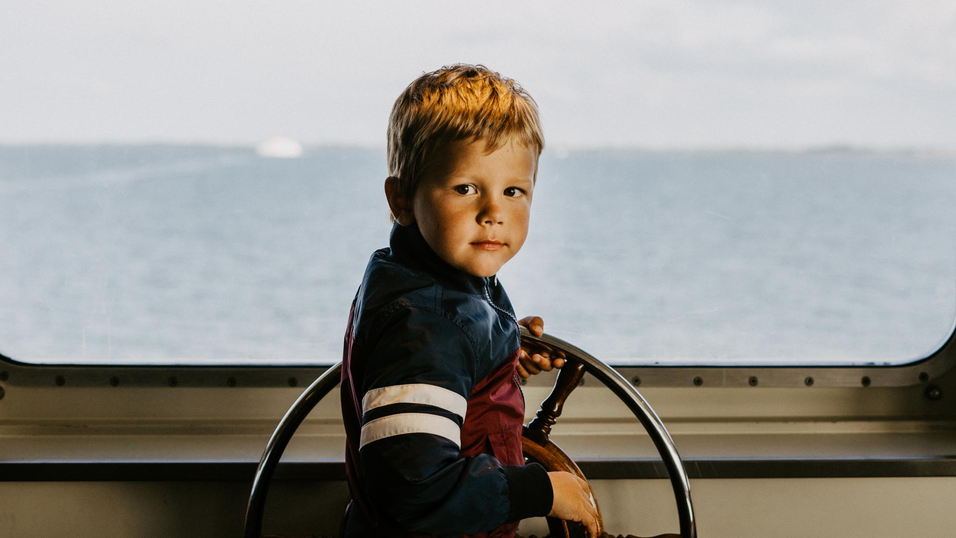 Ein Junge am Steuerrad eines Schiffs. Durch die Scheibe sieht man den Horizont und das Meer.