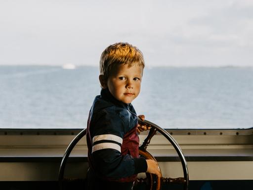 Ein Junge am Steuerrad eines Schiffs. Durch die Scheibe sieht man den Horizont und das Meer.