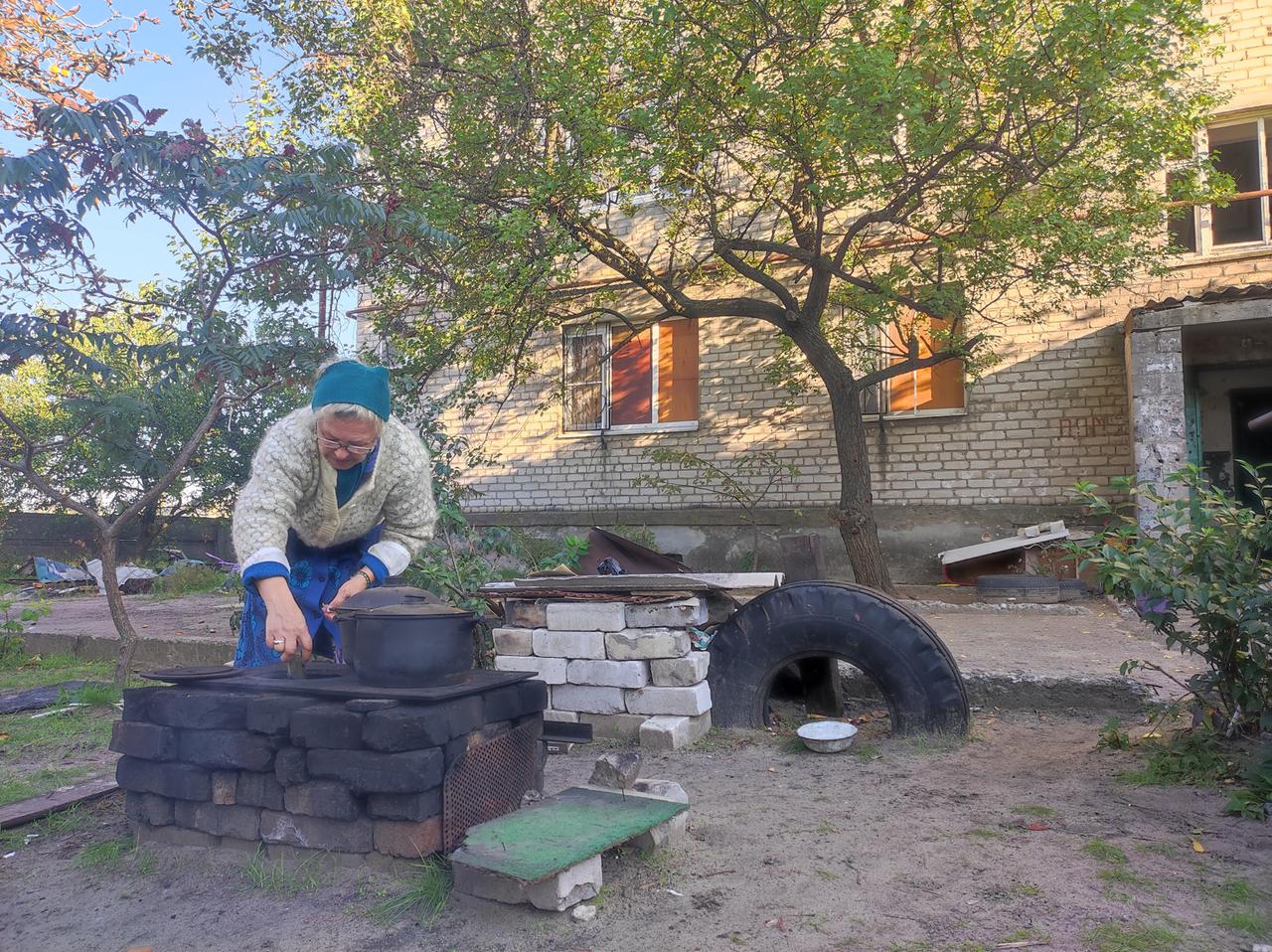Die Mutter von Maksim Surscha. Eine ältere Frau kocht auf einer offenen Feuerstelle im Freien.  