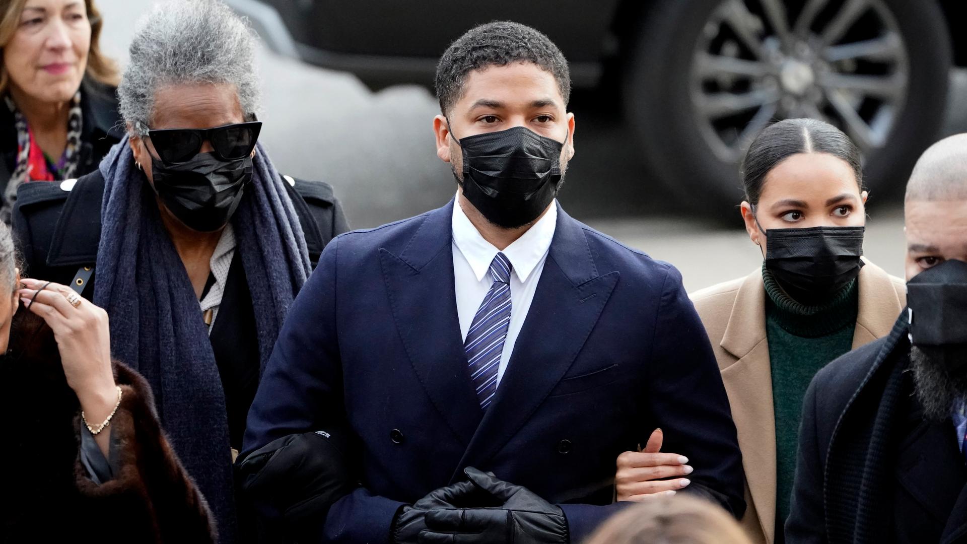 Smollett im blauen Jackett und mit Maske steht in der Mitte einer Menschengruppe auf einer Straße. Zwei Frauen sind bei ihm eingehakt. Smollett im blauen Jackett und mit Maske steht in der Mitte einer Menschengruppe auf einer Straße. Zwei Frauen sind bei ihm eingehakt.