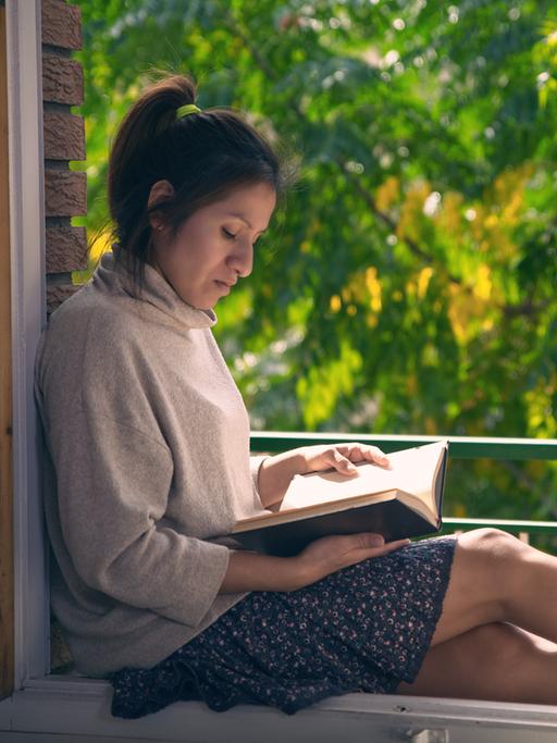 Eine Frau sitzt an einem geöffneten Fenster und liest ein Buch.