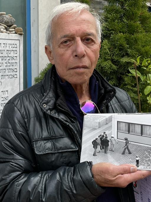 Shlomo Levy posiert mit einem historischen Foto neben der Gedenktafel im Olympischen Dorf in München für ein Foto.