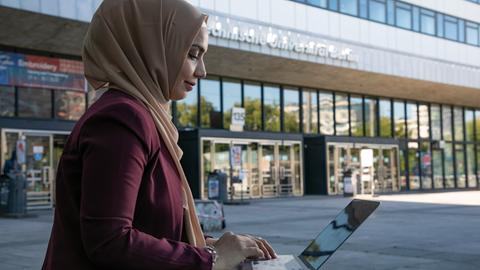 Eine junge Frau mit muslimischer Kopfbedeckung ist im Profil zu sehen. Sie sitzt im Freien vor einem Gebäude der Technischen Universität Berlin  und arbeitet an einem Laptop.