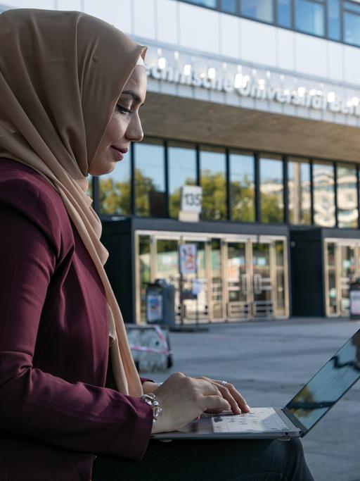 Eine junge Frau mit muslimischer Kopfbedeckung ist im Profil zu sehen. Sie sitzt im Freien vor einem Gebäude der Technischen Universität Berlin  und arbeitet an einem Laptop.