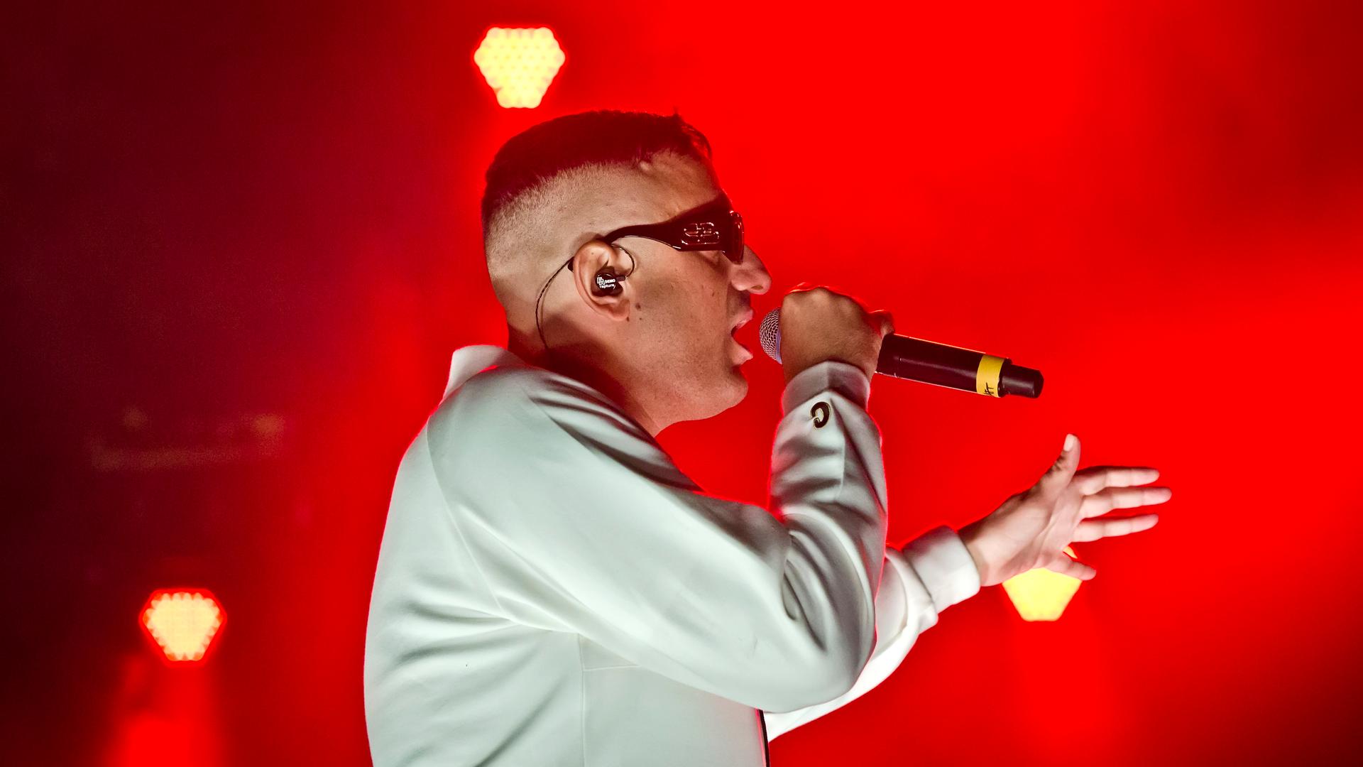Der Rapper Haftbefehl in einer weissen Jacke mit einer Sonnenbrille bei einem Auftritt auf einer rot erleuchteten Bühne.