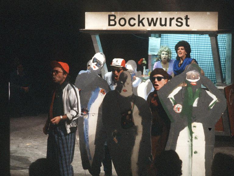 Szene des Rock-Musicals "Linie 1" im Berliner Grips-Theater an einem Bockwurststand, vor dem Menschen als Touristen verkleidet mit Sonnenbrille und Fotoapparaten sowie zusätzlichen Pappmaché-Figuren singen.