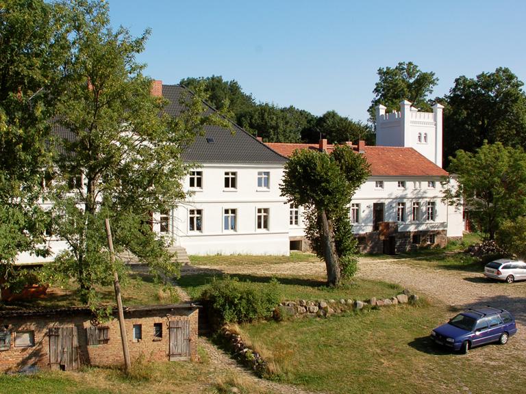 Aufnahme von Schloss Bröllin, im Vordergrund stehen links einige Bäume, das Gebäude mitsamt des mächtigen Turmes an der rechten Seite ist weiß angemalert. Im Hintergrund des Schlosses sind Baumkronen zu erkennen.