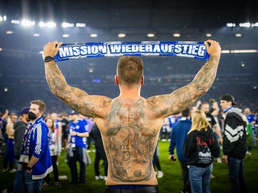 Ein Schalke Fan feiert den Aufstieg des FC Schalke 04 nach dem Spiel gegen St. Pauli am 07.05.2022. Er ist von hinten zu sehen und reckt einen Schal in zwischen beiden Armen in die Luft. Darauf steht: "Mission Wiederaufstieg".