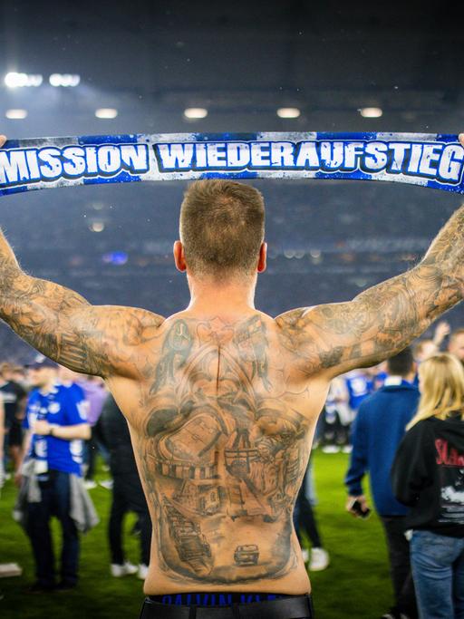 Ein Schalke Fan feiert den Aufstieg des FC Schalke 04 nach dem Spiel gegen St. Pauli am 07.05.2022. Er ist von hinten zu sehen und reckt einen Schal in zwischen beiden Armen in die Luft. Darauf steht: "Mission Wiederaufstieg".