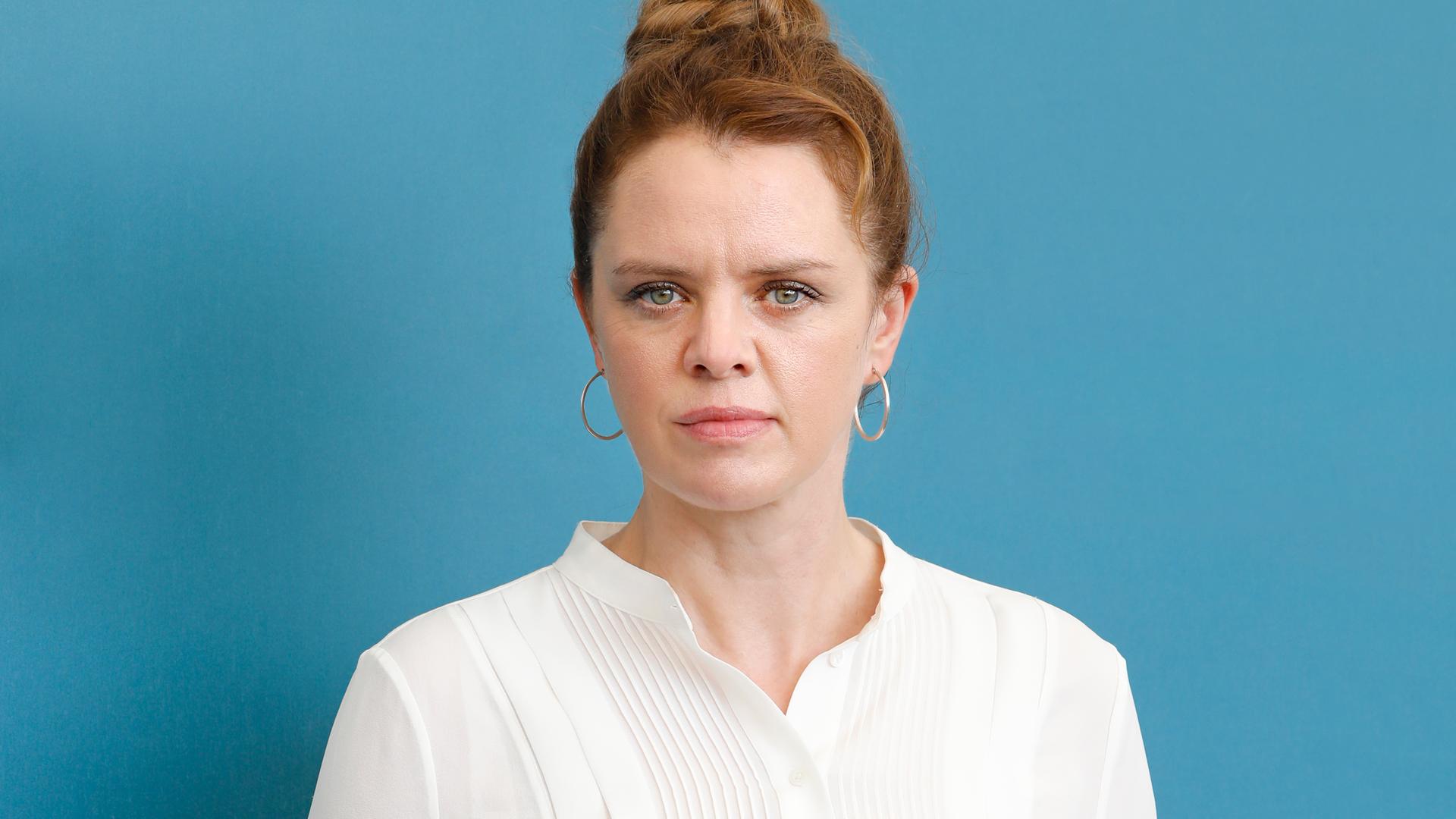 Julia von Heinz steht mit weißer Bluse und schwarzer Hose vor einem hellblauen Hintergrund und schaut in die Kamera Julia von Heinz steht mit weißer Bluse und schwarzer Hose vor einem hellblauen Hintergrund und schaut in die Kamera