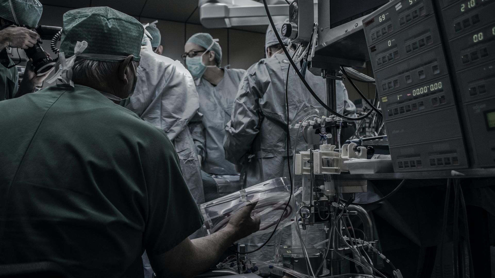 Ärzte in OP-Kitteln drängen sich im OP Saal, zu sehen ist außerdem eine Maschine mit vielen Kabeln.