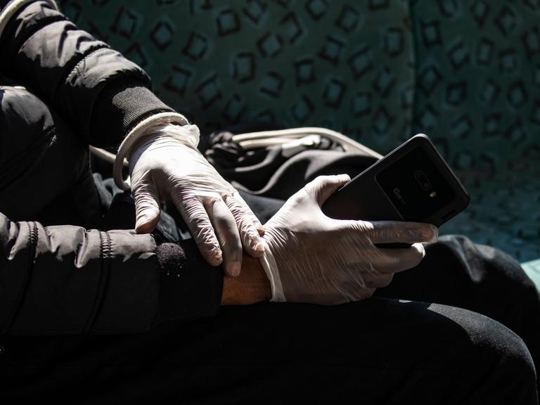 Eine Person in schwarzer Jacke hält sein Smartphone mit Schutzhandschuhen in der S-Bahn am Alexanderplatz in Berlin.