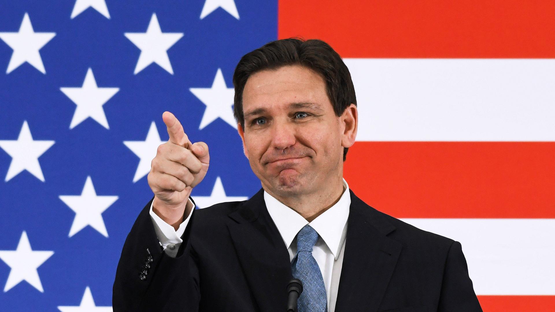 Der Gouverneur von Florida ist Ron DeSantis. Er steht vor einer US-Flagge und zeigt nach vorne.