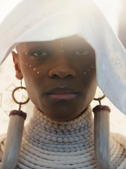 Letitia Wright als Shuri in "Black Panther: Wakanda Forever". Sie trägt einen weißen Schleier auf dem Kopf, übergroße Ohrringe und blickt demonstrativ in die Kamera.