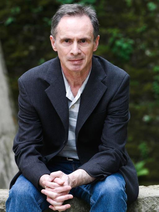 Der serbische Schriftsteller David Albahari sitzt in blauer Jeans, hellem Shirt und dunklem Sakko auf einer Steinmauer vor grünem Hintergrund.