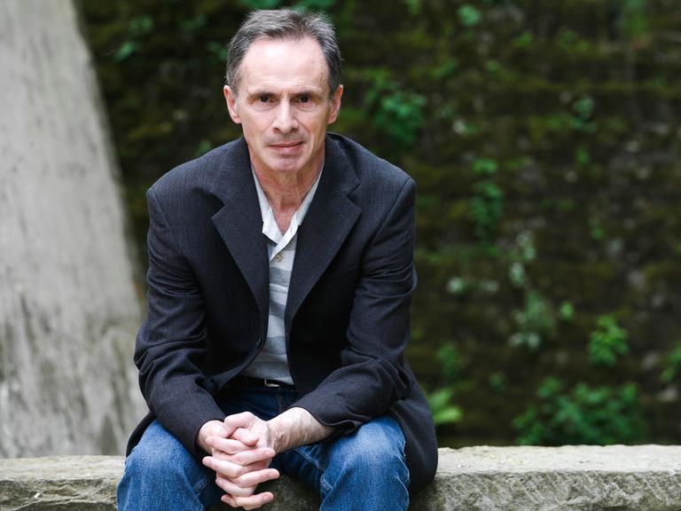 Der serbische Schriftsteller David Albahari sitzt in blauer Jeans, hellem Shirt und dunklem Sakko auf einer Steinmauer vor grünem Hintergrund.