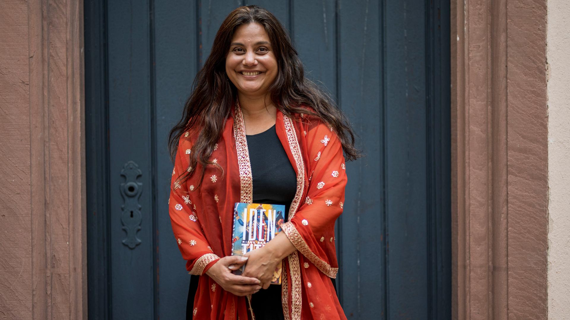 Porträt von Mithu Sanyal, die eine rot-orangene Jacke trägt und mit einem Buch in der Hand lachend vor einer schwarzen Tür steht. Porträt von Mithu Sanyal, die eine rot-orangene Jacke trägt und mit einem Buch in der Hand lachend vor einer schwarzen Tür steht.