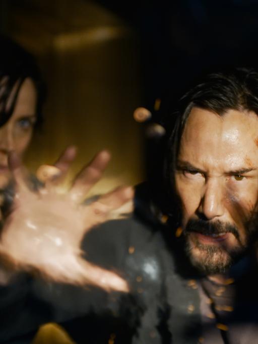 Keanu Reeves (Neo) und Carrie-Ann Moss (Trinity) in einer Szene des Films "Matrix Resurrections". Neo streckt die Hände einem unbekannten Gegner entgegen, Trinity blickt im Hintergrund furchtsam.