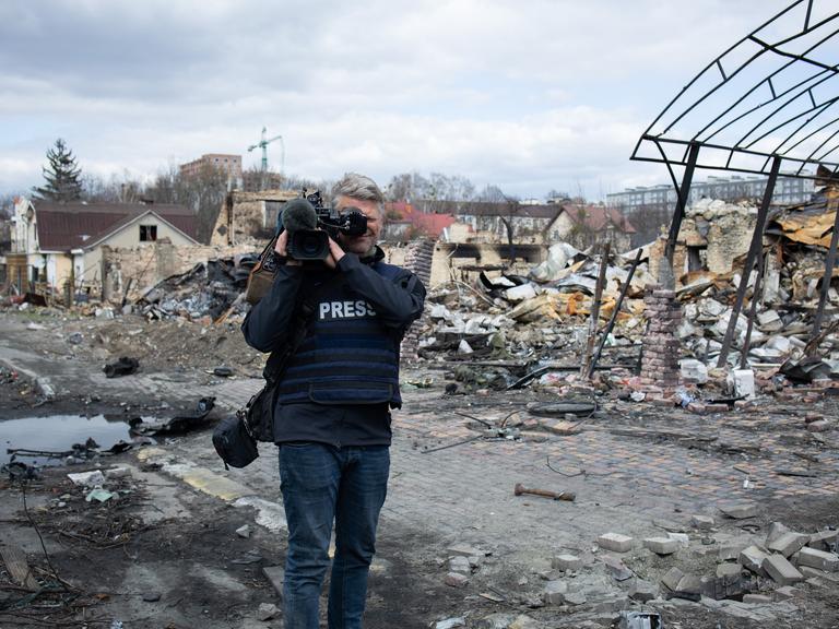 Ein Journalist mit Kamera und einer Jacke mit der Aufschrift "PRESS" steht vor zerstörten Gebäuden.